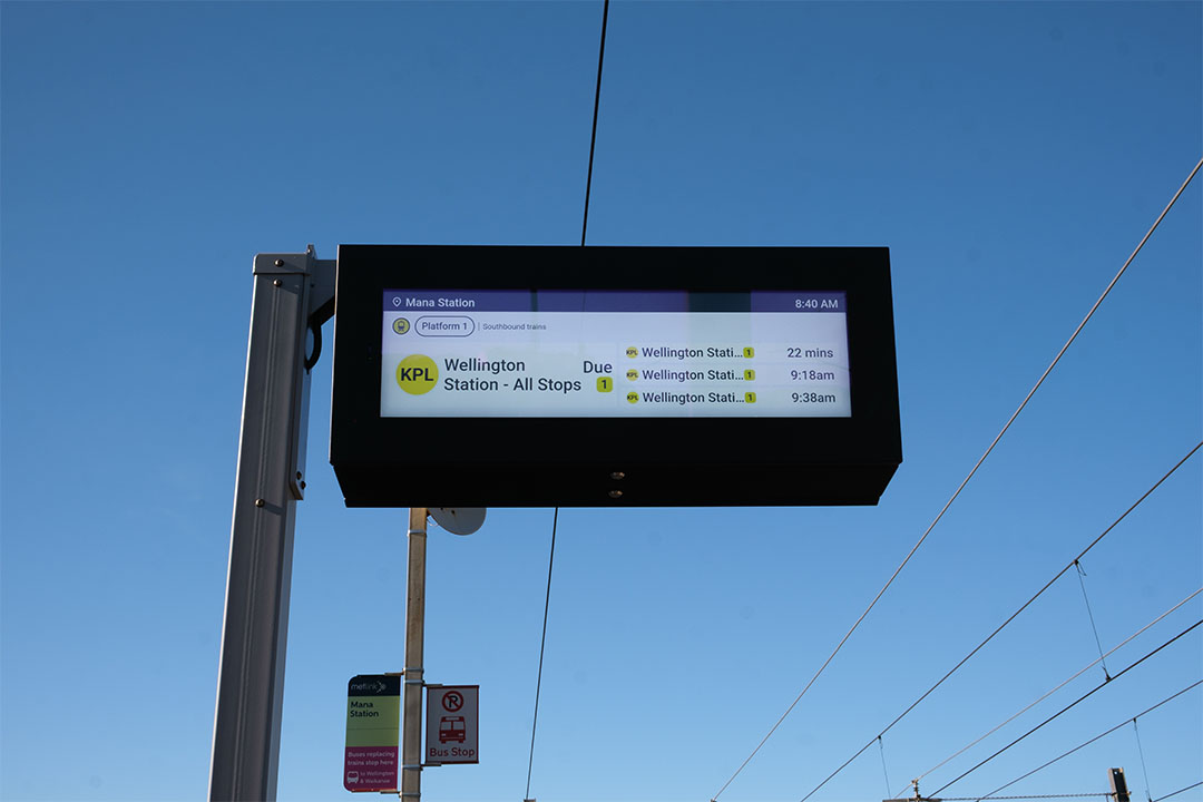 digital screen at mana station showing train arrival times with bus stop sign in the background