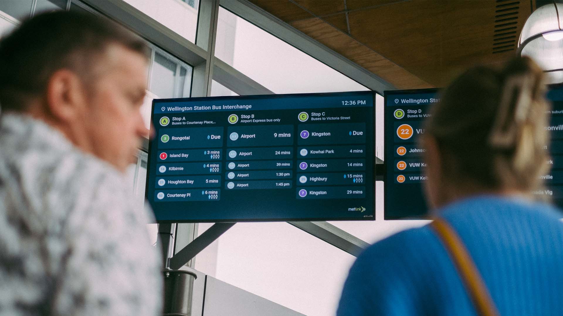 two commuters looking up at Wellington Station Bus Interchange information across two digital screens
