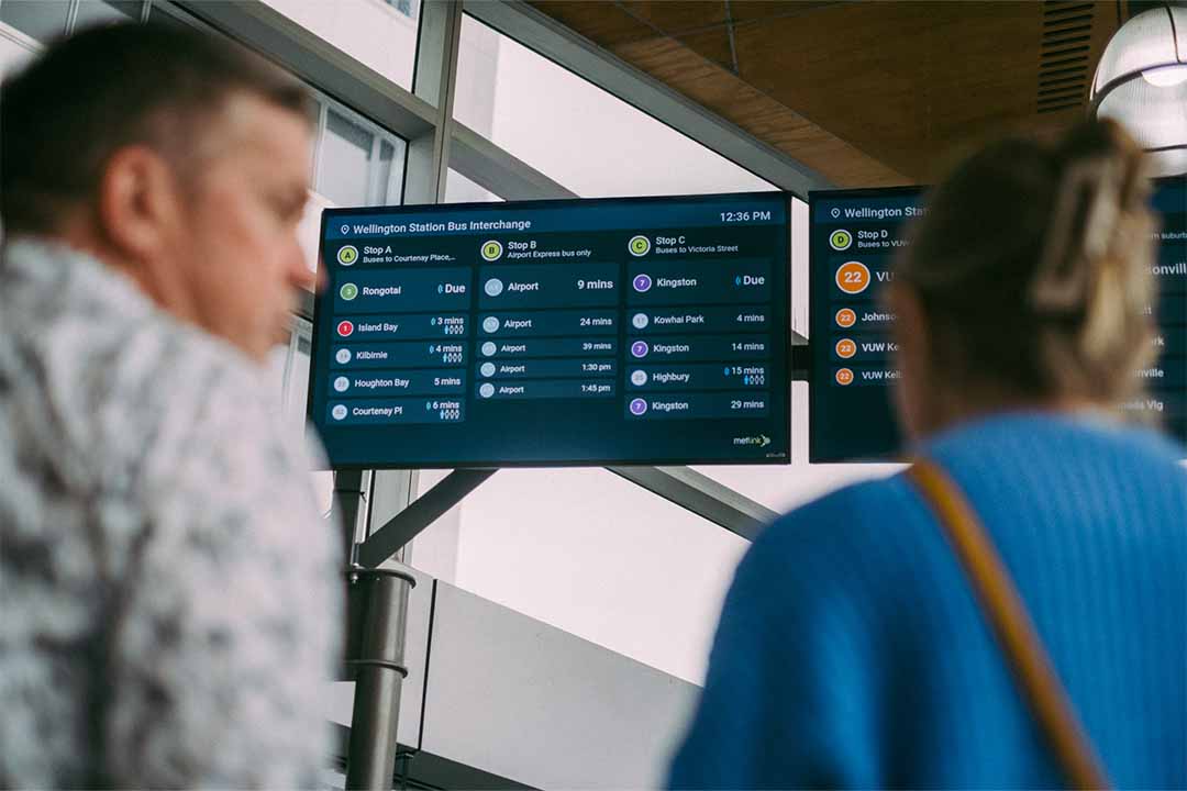 two commuters looking up at a digital screen displaying bus information